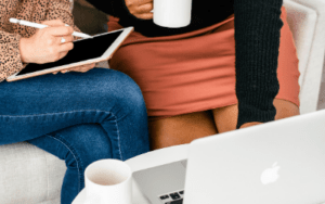 two women in front of a laptop with cups a coffee, one in jeans one in an orange skirt